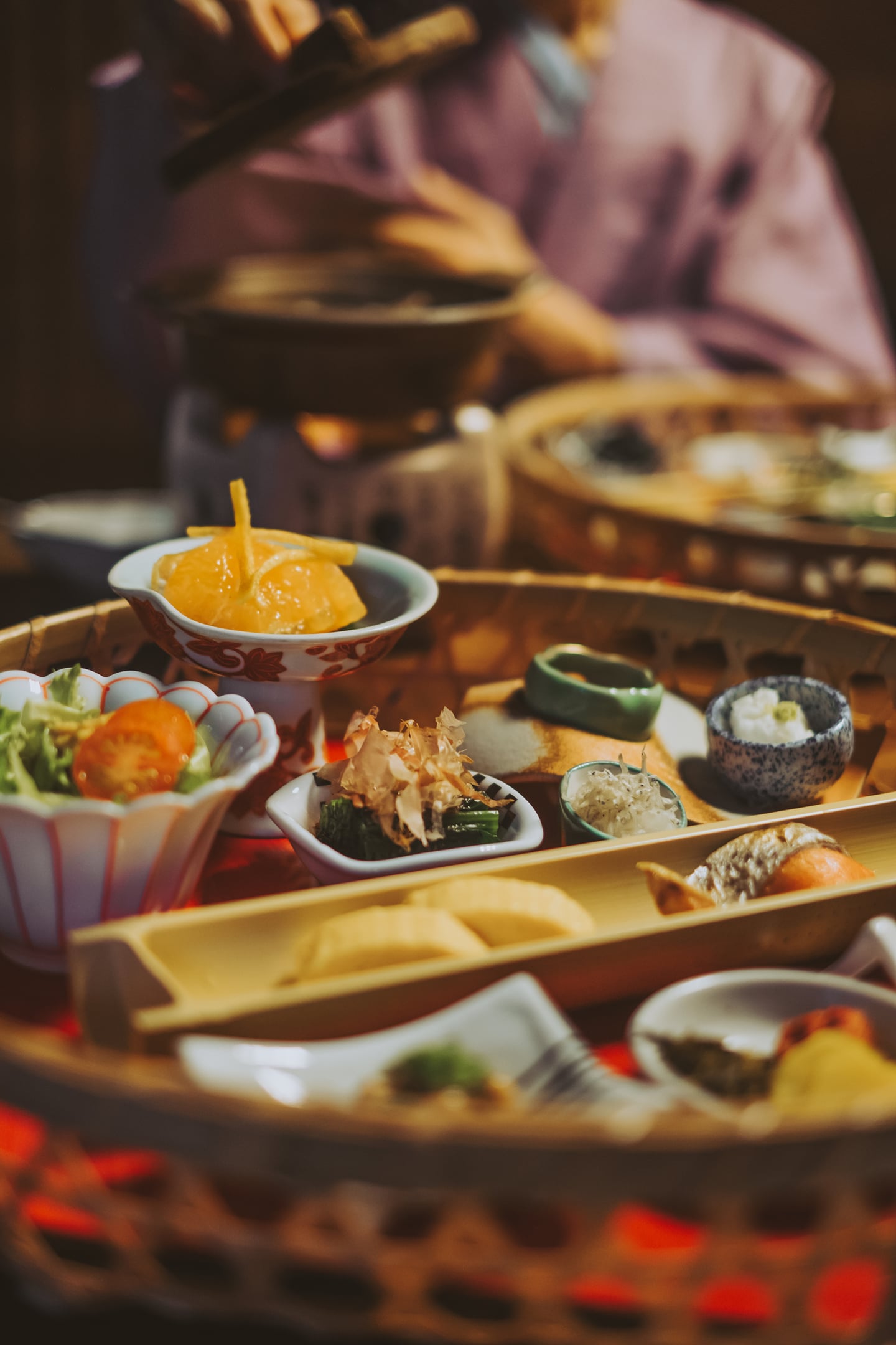 전통적인 대나무 바구니에 담긴 다채로운 일본식 정찬 요리와 유카타를 입은 인물이 어우러진 따뜻한 분위기의 식사 장면입니다.