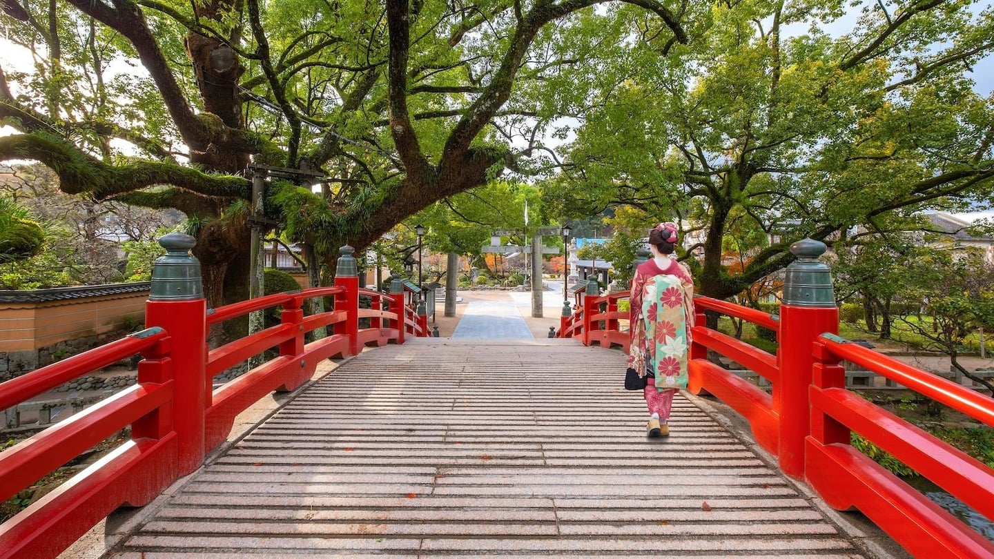 붉은 난간이 돋보이는 일본 전통 다리 위에서 화려한 꽃무늬 기모노를 입고 산책하는 여성의 뒷모습과 푸른 나무가 어우러진 풍경입니다.