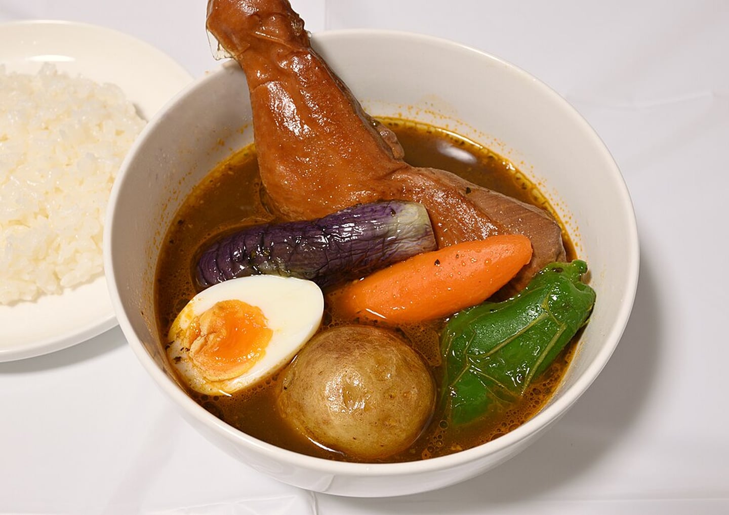 진한 국물에 큼직한 닭다리와 알록달록한 채소 토핑이 어우러진 먹음직스러운 스프카레와 흰 쌀밥이 놓인 모습입니다.