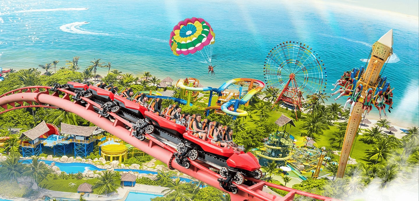 에메랄드빛 바다 옆으로 펼쳐진 대형 테마파크에서 롤러코스터와 관람차를 즐기는 사람들과 하늘 위 알록달록한 패러세일링이 어우러진 역동적인 풍경입니다.