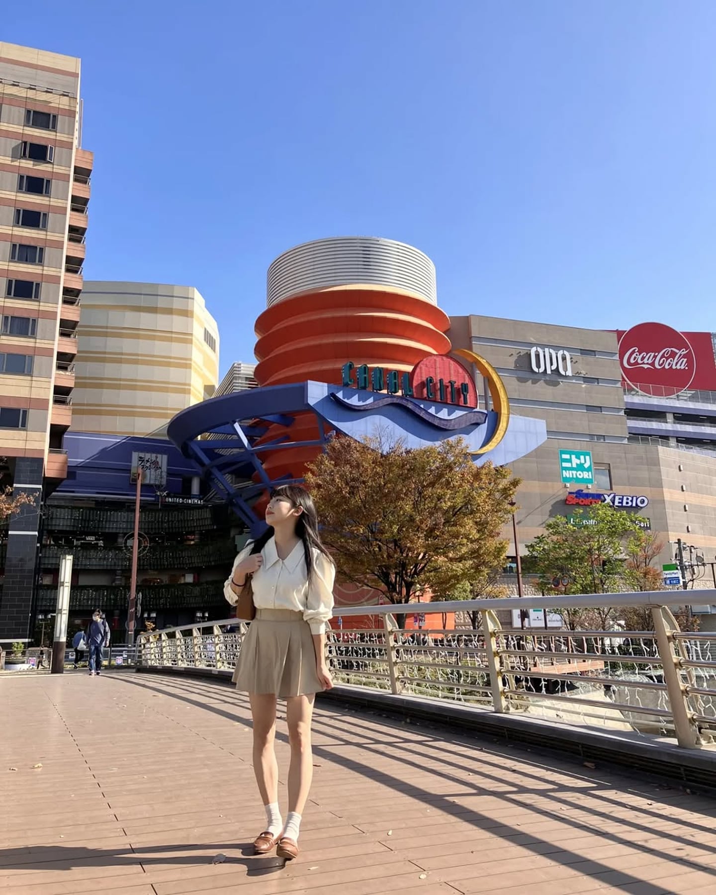 후쿠오카 캐널시티의 상징적인 붉은색 조형물과 현대적인 쇼핑몰 건물을 배경으로 베이지색 스커트를 입은 여성이 다리 위에서 여유롭게 산책하는 모습입니다.