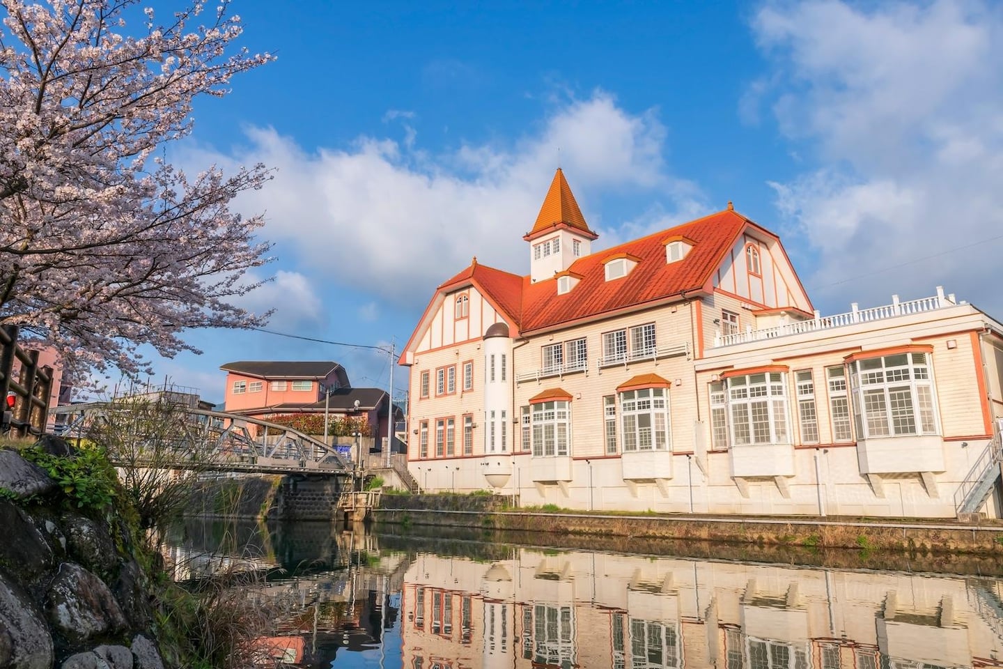 푸른 하늘과 뭉게구름 아래 강가에 위치한 주황색 지붕의 유럽풍 건물과 분홍빛 벚꽃이 만발한 아름다운 봄의 수변 경관입니다.
