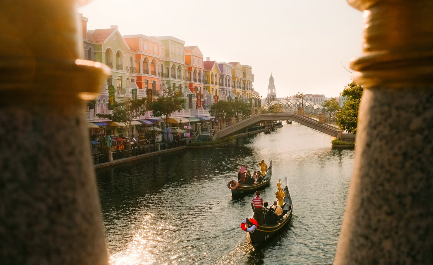 석양이 비치는 운하 위로 곤돌라가 지나가고 파스텔 톤의 화려한 건물들과 아치형 다리가 어우러진 이국적인 테마파크의 전경입니다.