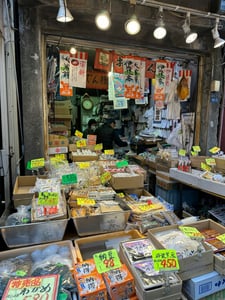 일본 전통 시장의 식료품점 앞에 어묵과 나또 등 다양한 식재료가 담긴 상자들이 층층이 쌓여 있고 상인들이 일하는 활기찬 풍경입니다.