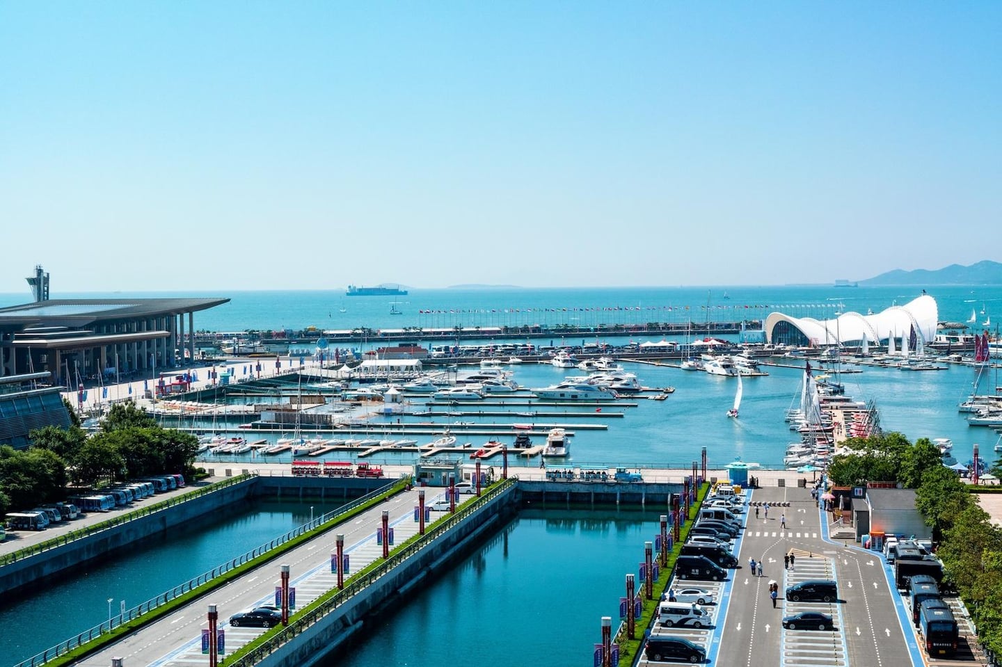 푸른 바다 위로 하얀 요트들이 가득한 마리나 항구와 곡선미가 돋보이는 현대적인 건축물이 조화를 이루는 맑은 날의 해안가 풍경입니다.