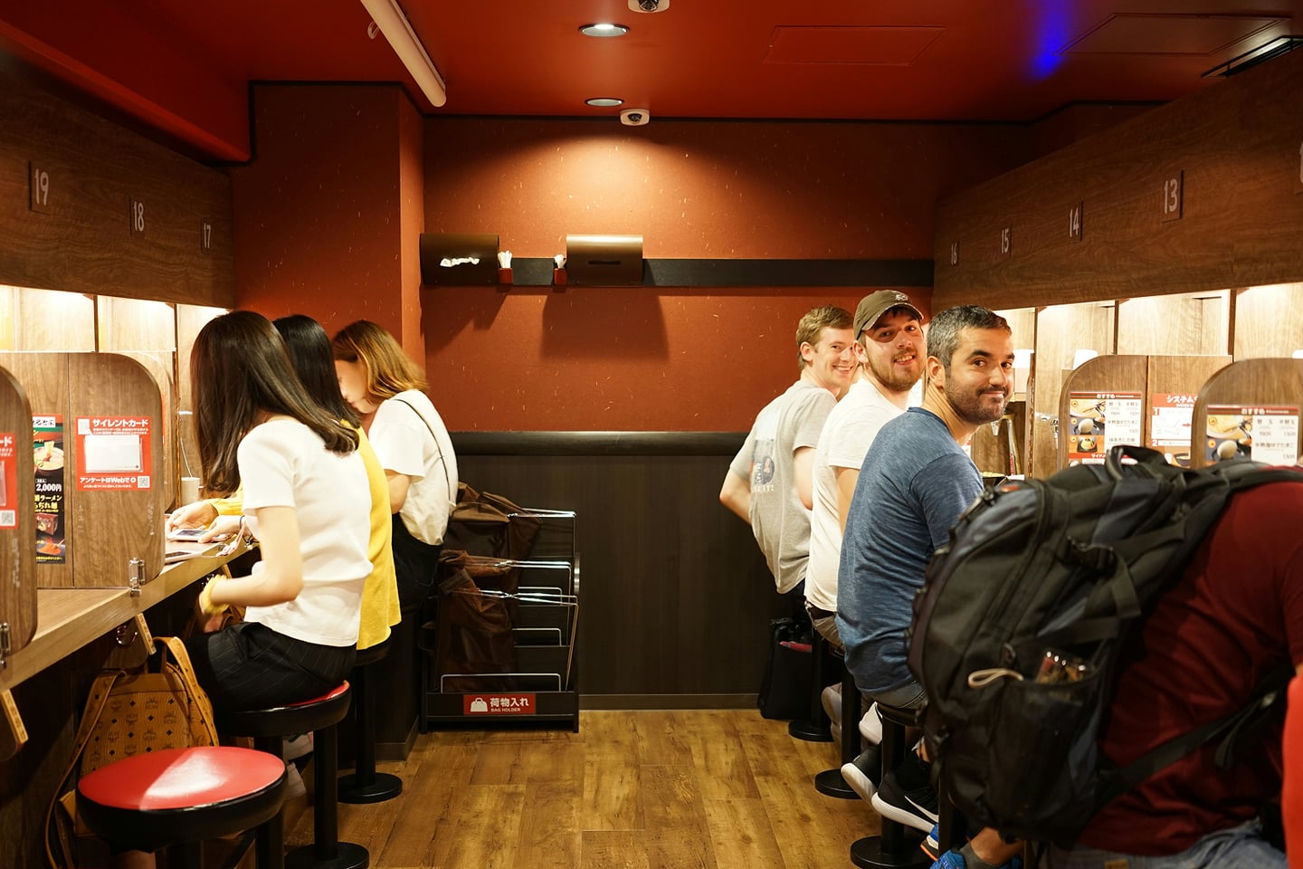 칸막이가 설치된 일본 라멘 식당의 1인용 카운터 좌석에 나란히 앉아 식사를 기다리는 사람들의 모습입니다.