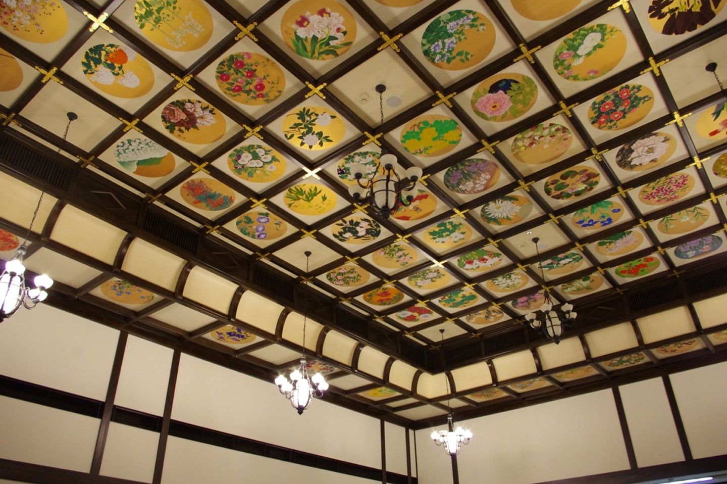 격자형 천장을 가득 채운 다채로운 꽃 그림들과 은은한 빛을 내는 고전적인 샹들리에가 어우러진 일본 사찰 혹은 전통 가옥의 내부 전경입니다.