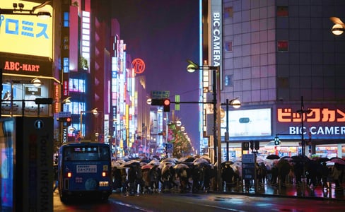 비 오는 밤 일본 신주쿠 거리에서 우산을 쓴 인파가 횡단보도를 건너고 있으며, 주변의 화려한 네온사인과 빅카메라 간판이 젖은 노면에 반사되어 반짝이는 풍경입니다.