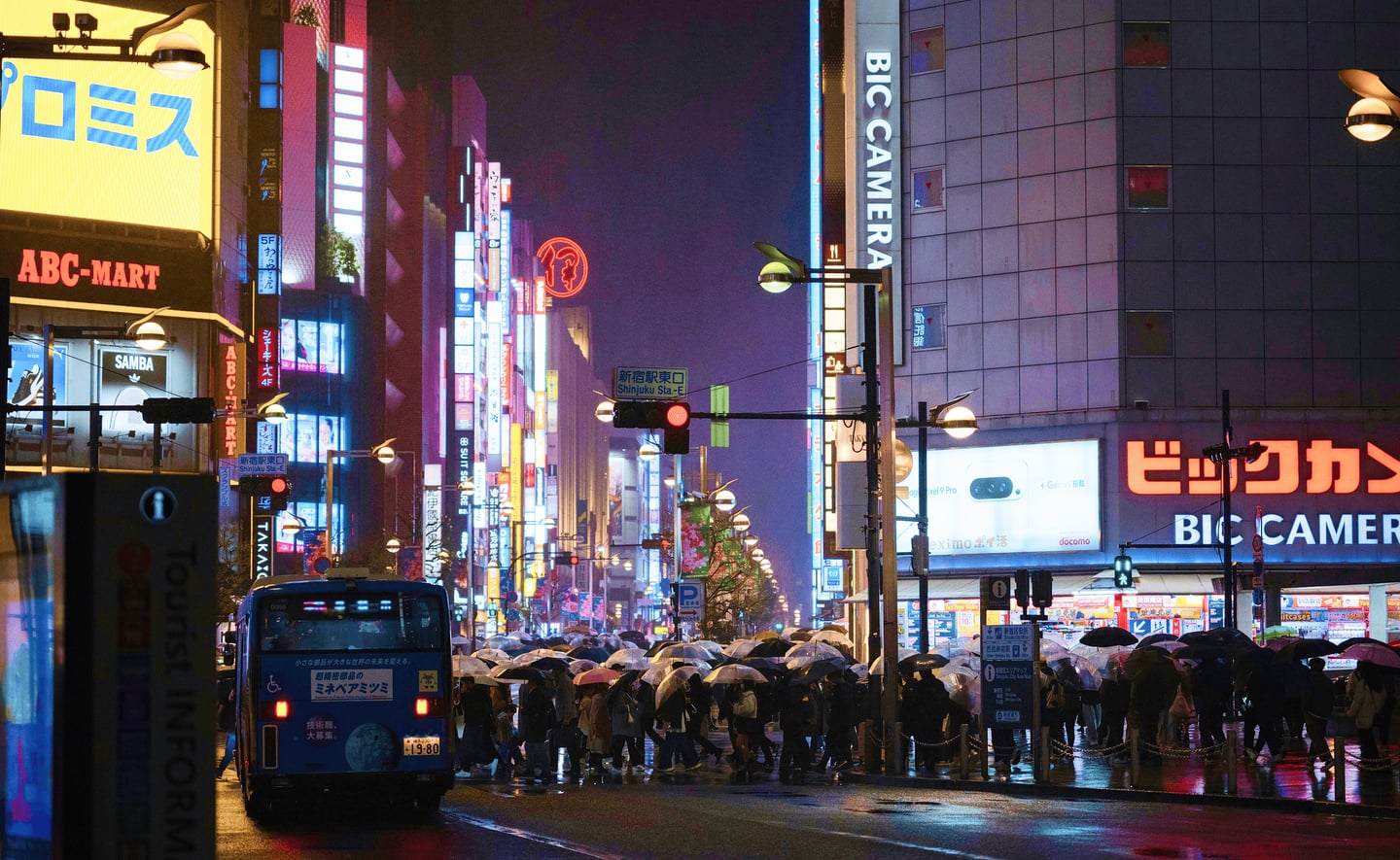 비 오는 밤 일본 신주쿠 거리에서 우산을 쓴 인파가 횡단보도를 건너고 있으며, 주변의 화려한 네온사인과 빅카메라 간판이 젖은 노면에 반사되어 반짝이는 풍경입니다.