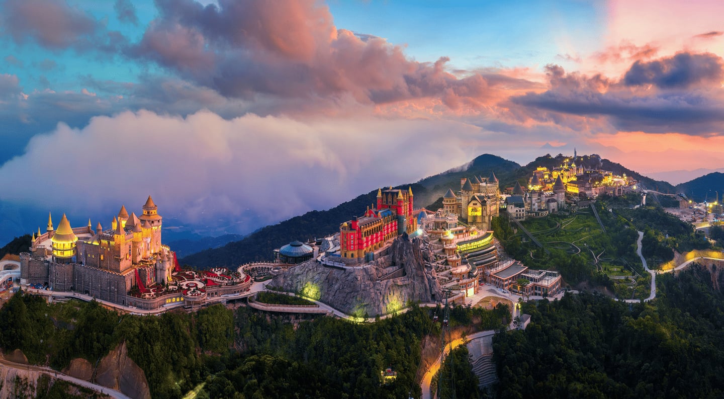 노을이 지는 하늘 아래 구름과 안개로 둘러싸인 바나힐의 화려한 성들과 유럽풍 건축물들이 산 정상에 펼쳐진 환상적인 야경 모습입니다.