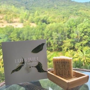 울창한 산을 배경으로 야외 테이블 위에 놓인 LUI & JULI 천연 비누와 카탈로그가 자연과 어우러져 평화롭고 고급스러운 느낌을 줍니다.