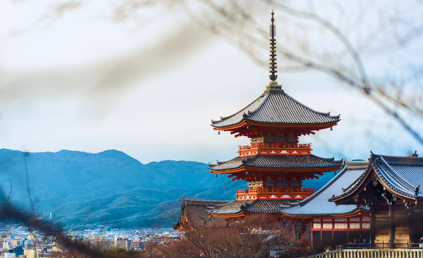 푸른 산과 도시 전경이 내려다보이는 언덕 위로 화려한 주황색 기둥과 기와지붕이 조화를 이룬 일본 교토 기요미즈데라의 3층 탑 전경입니다.