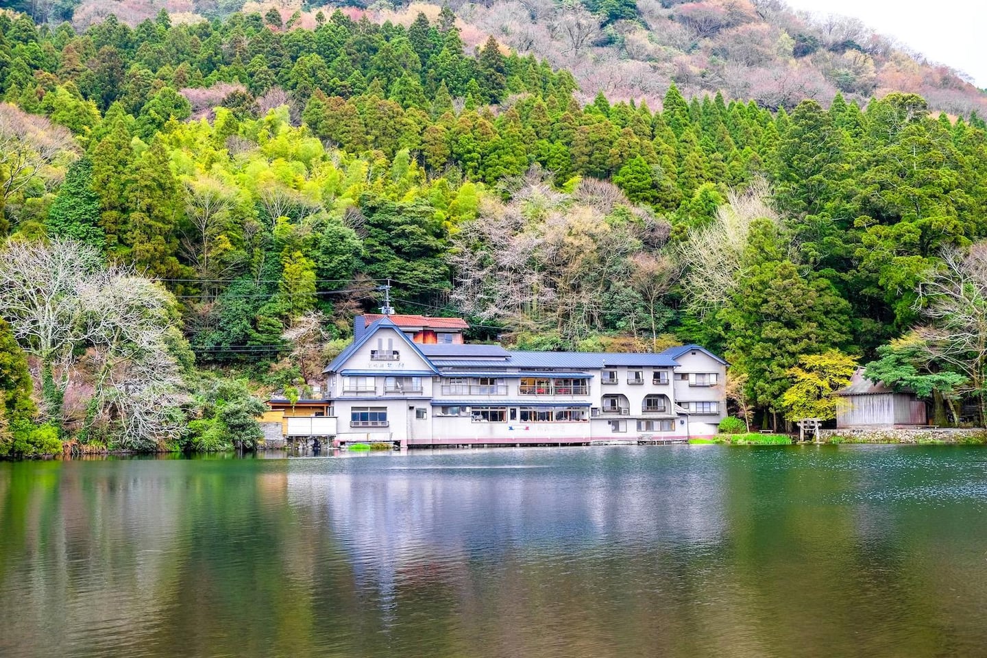 푸른 나무가 가득한 산을 배경으로 호수 바로 옆에 자리 잡은 회색빛 2층 건물이 수면에 비치는 평온한 자연 경관입니다.