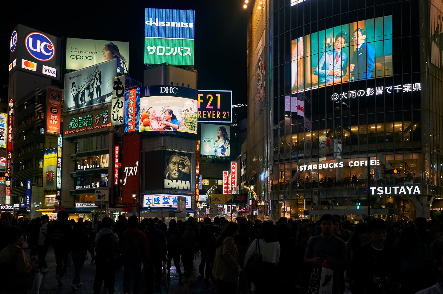 화려한 네온사인과 대형 전광판 광고가 가득한 도쿄 시부야 거리의 밤 풍경 속에서 수많은 사람들이 횡단보도를 건너는 모습입니다.