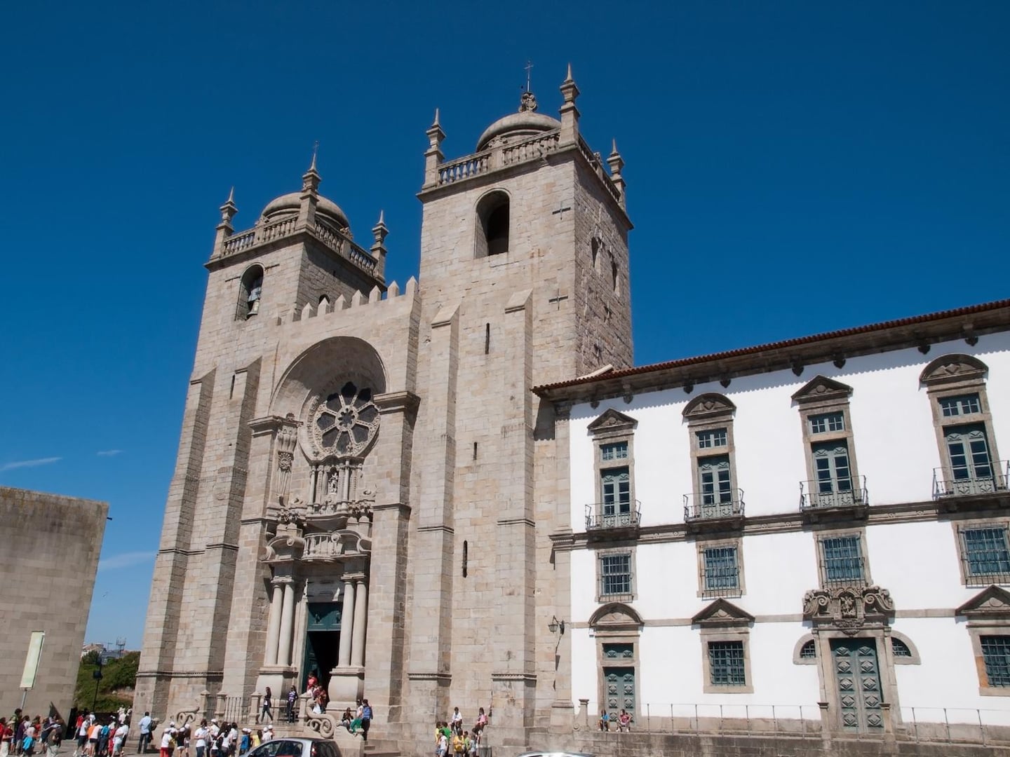 포르투갈 포르투 대성당의 거대한 석조 타워와 정교한 장미창 장식, 그리고 옆으로 이어진 고전적인 양식의 흰색 벽면 건물이 대비를 이루는 풍경입니다.
