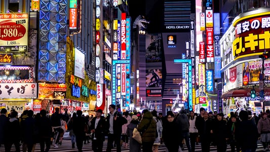 화려한 네온사인과 고질라 조형물이 눈에 띄는 도쿄 신주쿠 가부키초 거리에서 밤늦게 횡단보도를 건너는 수많은 사람들의 모습입니다.