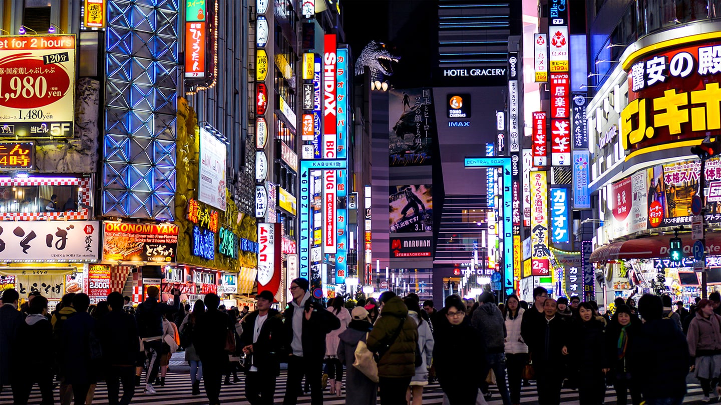 화려한 네온사인과 고질라 조형물이 눈에 띄는 도쿄 신주쿠 가부키초 거리에서 밤늦게 횡단보도를 건너는 수많은 사람들의 모습입니다.