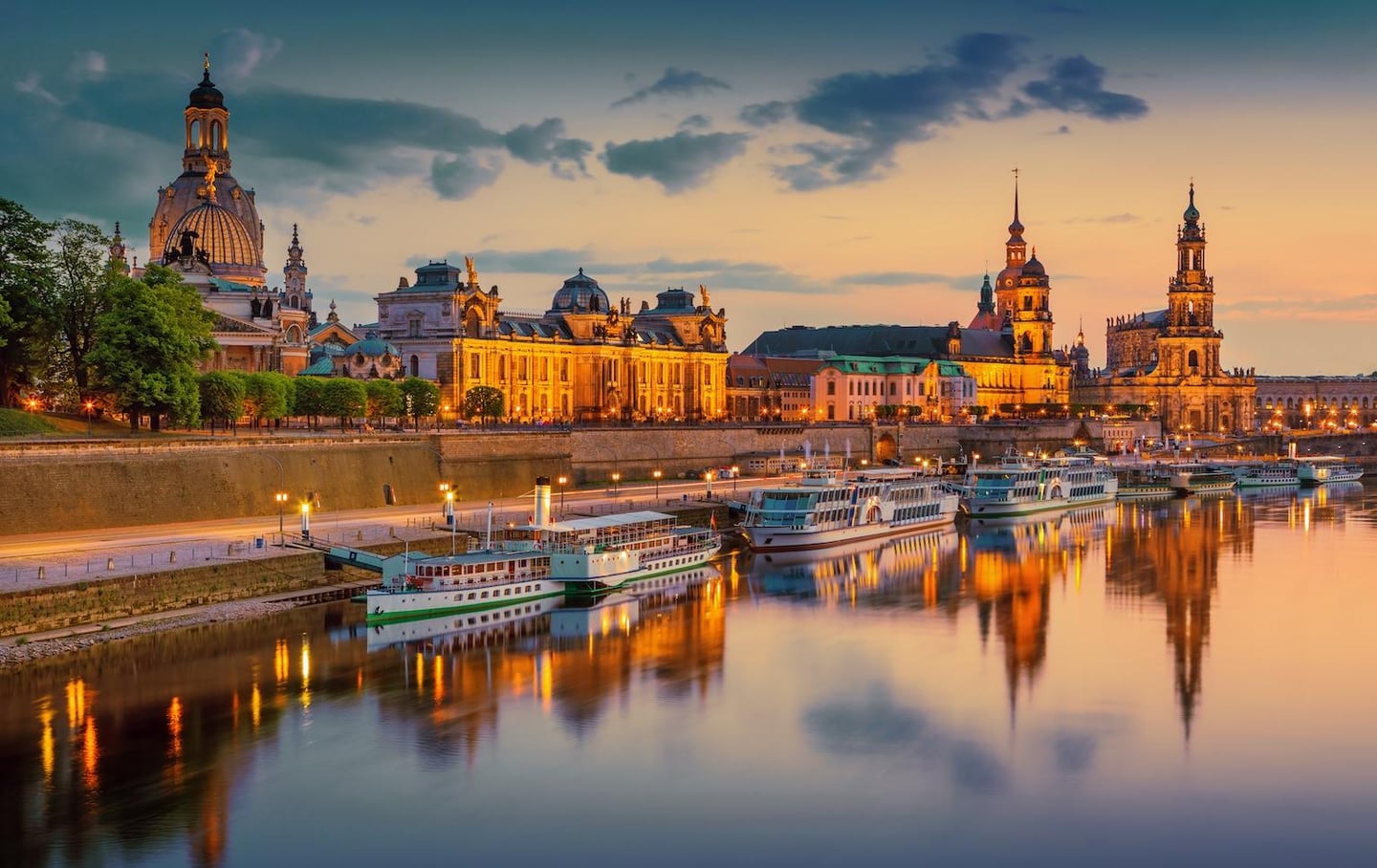 황금빛 조명이 켜진 드레스덴의 역사적인 건축물들과 엘베 강 위에 떠 있는 유람선들이 노을 진 하늘과 어우러진 평화로운 저녁 풍경입니다.