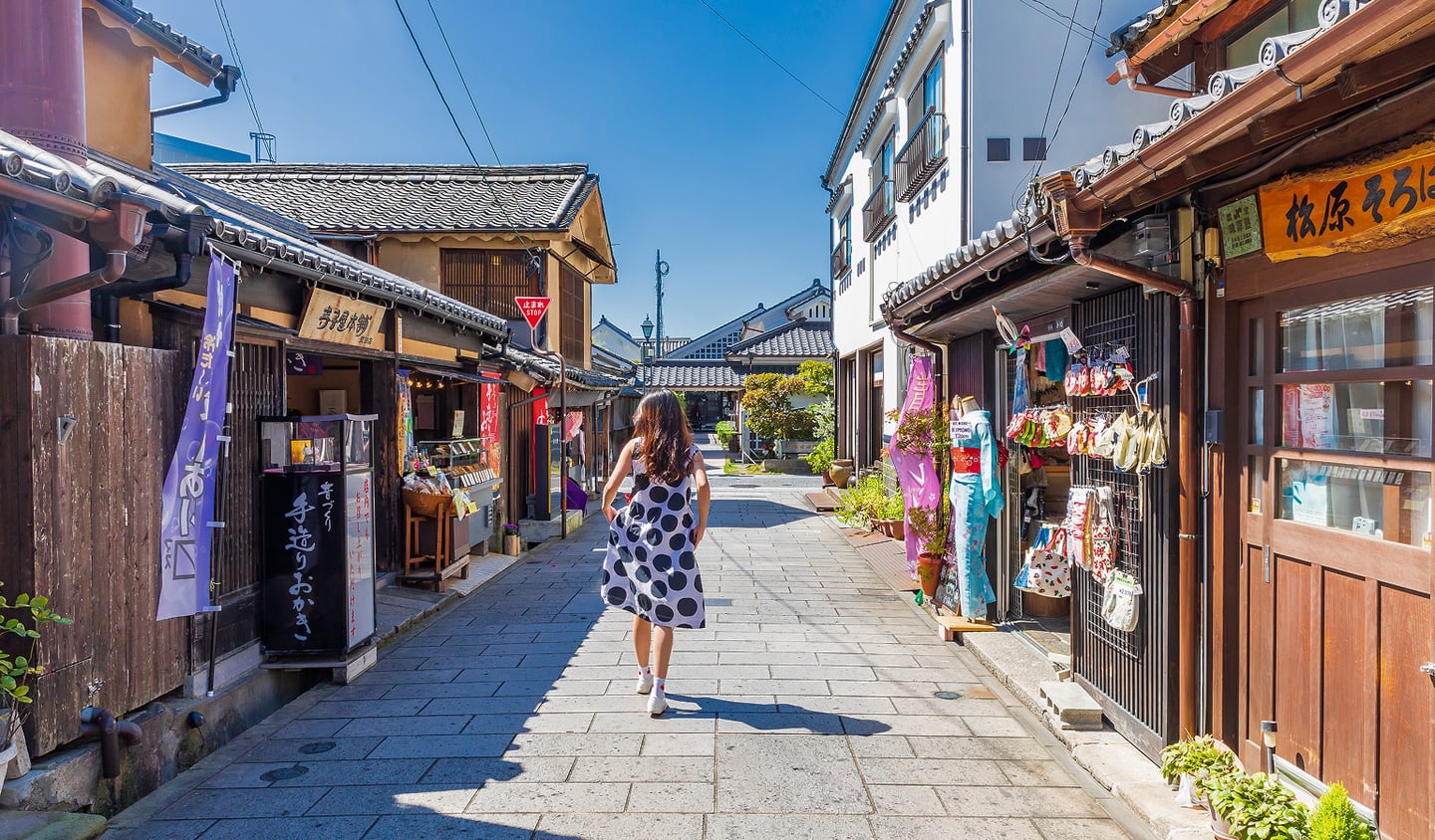 일본의 고풍스러운 전통 가옥과 상점들이 늘어선 돌바닥 골목길에서 도트 무늬 드레스를 입은 여행객이 여유롭게 산책을 즐기는 모습입니다.