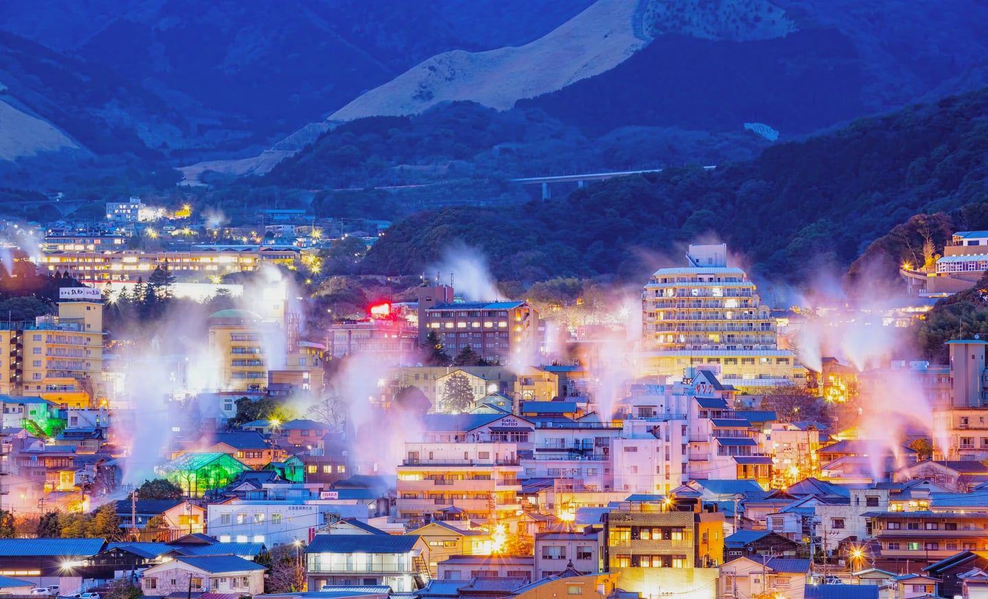 어스름한 저녁 푸른 산 아래로 온천 수증기가 피어오르는 일본 벳푸 마을의 화려한 야경과 평화로운 풍경입니다.