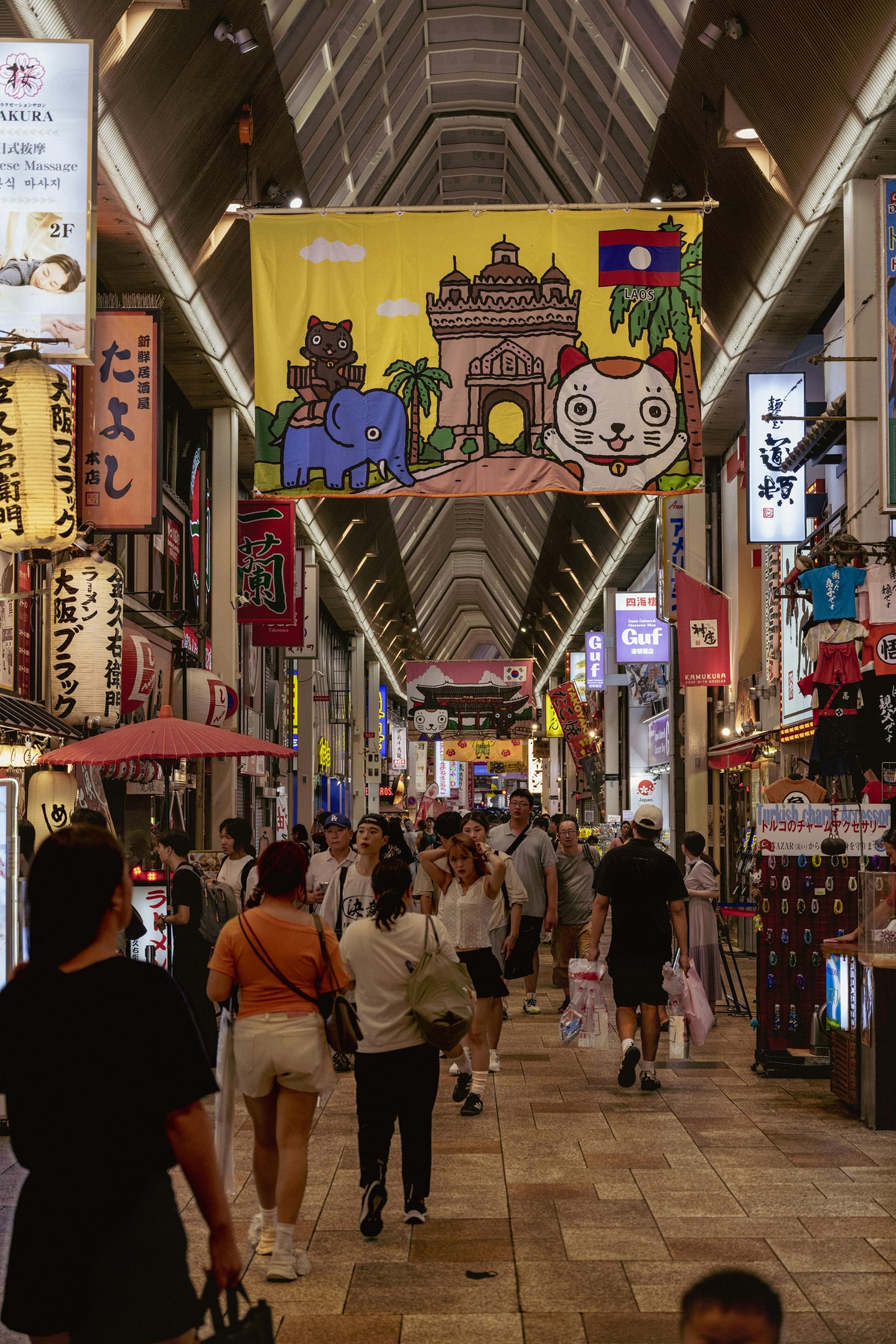 일본 오사카 도톤보리의 활기찬 아케이드 상점가에서 관광객들이 화려한 캐릭터 현수막과 일본어 간판 사이를 걷고 있는 모습입니다.