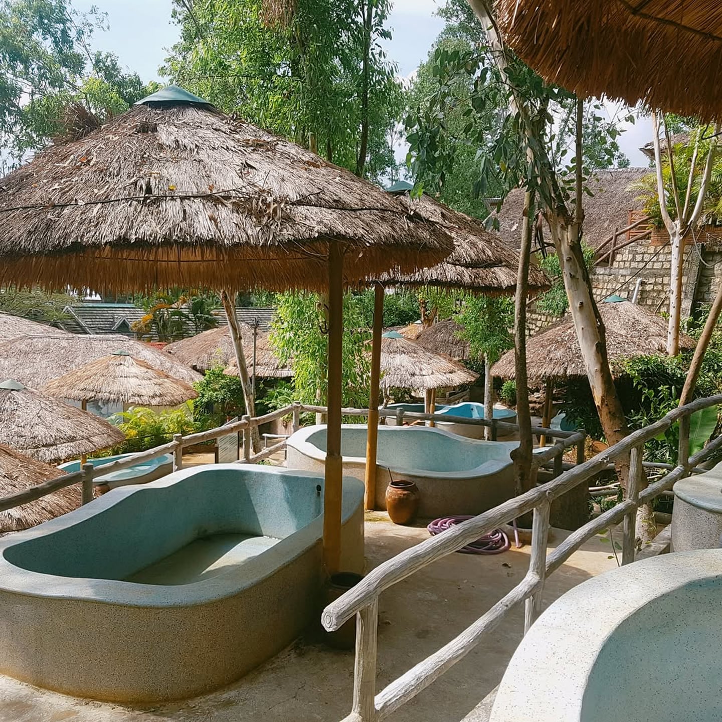 푸른 숲속에 자리 잡은 이국적인 초가 파라솔 아래로 둥근 곡선의 야외 욕조들이 늘어서 있는 평온한 스파 리조트의 전경입니다.