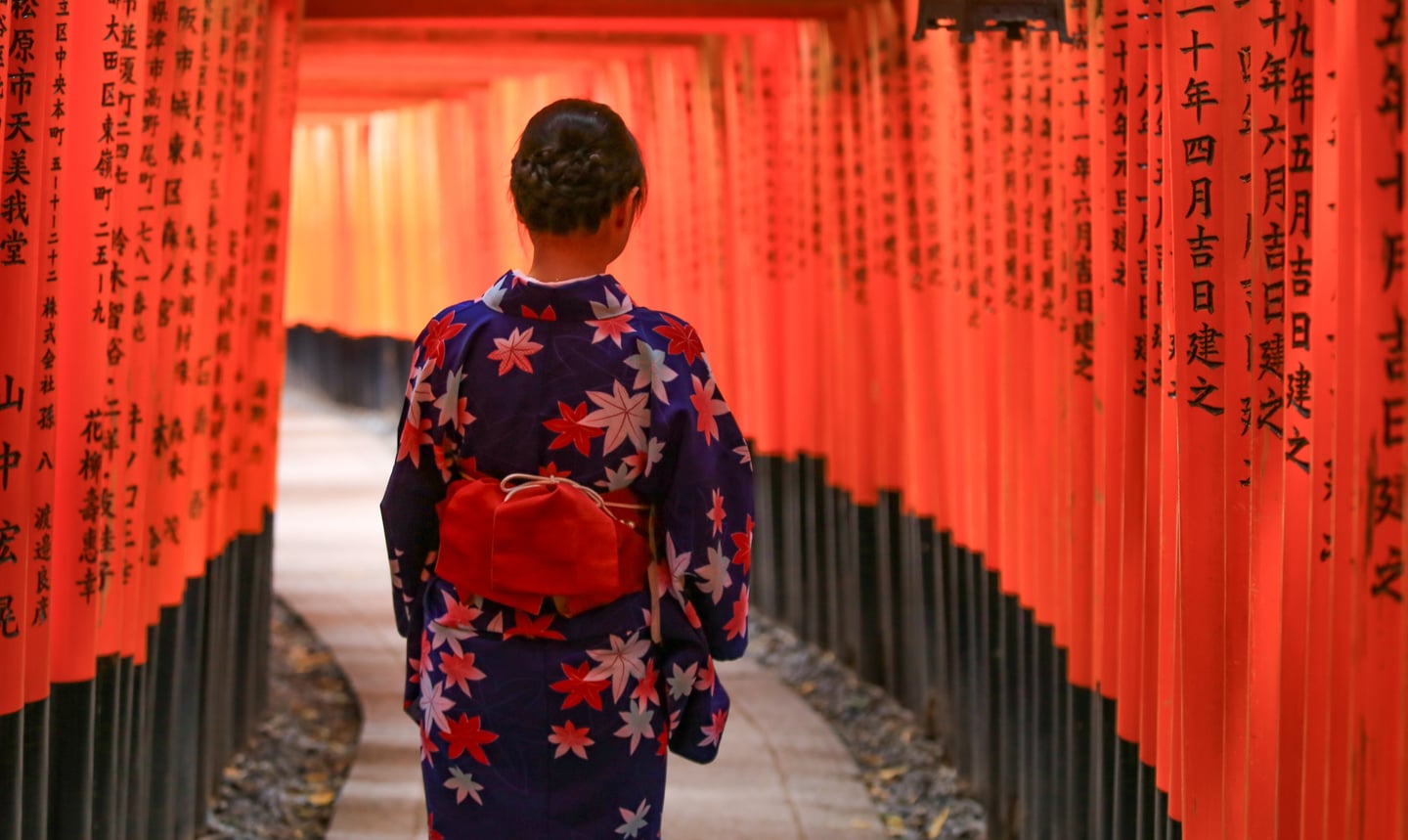 일본 교토 후시미 이나리 신사의 붉은 도리이 터널 아래에서 화려한 꽃무늬 기모노를 입고 산책하는 여성의 뒷모습이 평화로운 분위기를 자아냅니다.