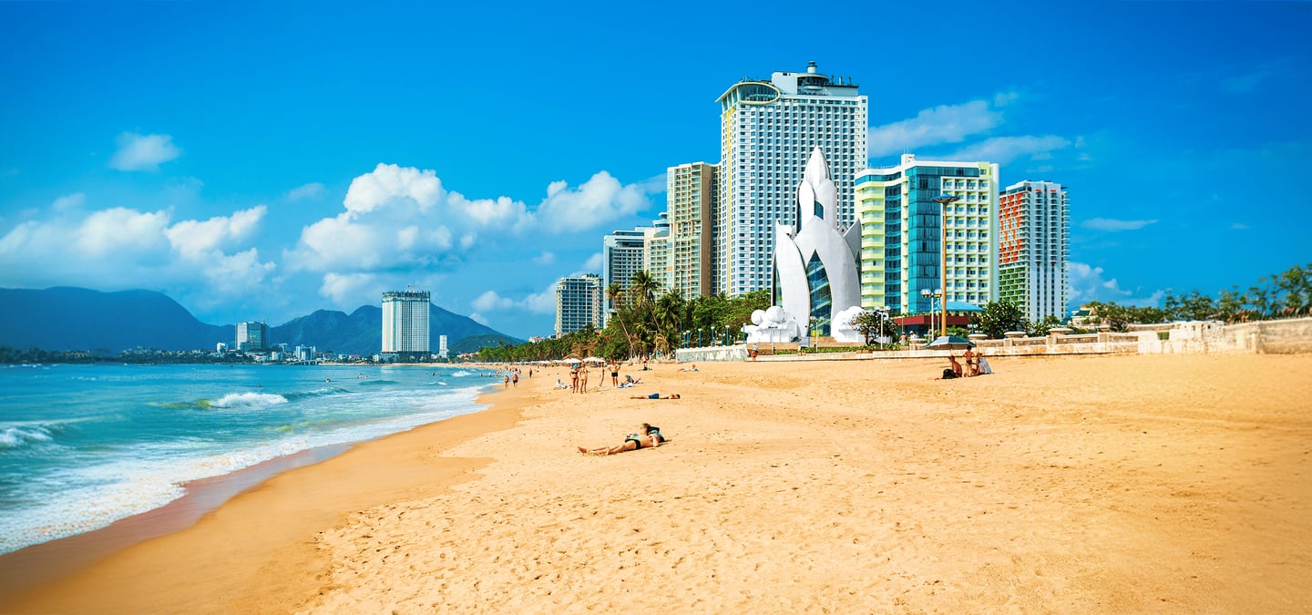 맑고 푸른 바다와 황금빛 모래사장이 펼쳐진 베트남 나트랑 해변을 배경으로 독특한 흰색 조형물과 고층 호텔들이 늘어선 이국적인 풍경입니다.