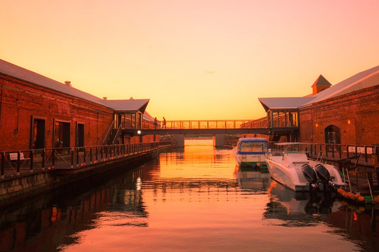해 질 녘 오렌지빛으로 물든 운하를 따라 늘어선 붉은 벽돌 창고와 정박한 보트들이 어우러진 이국적인 항구 도시의 저녁 풍경입니다.