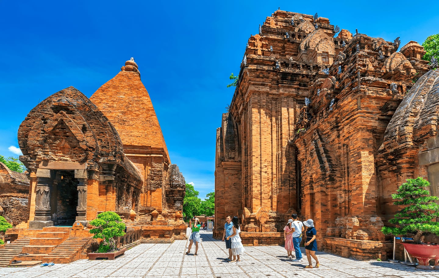 베트남 나트랑의 포나가르 첨탑에서 관광객들이 푸른 하늘을 배경으로 붉은 벽돌의 고대 유적지를 관람하며 산책하는 풍경입니다.