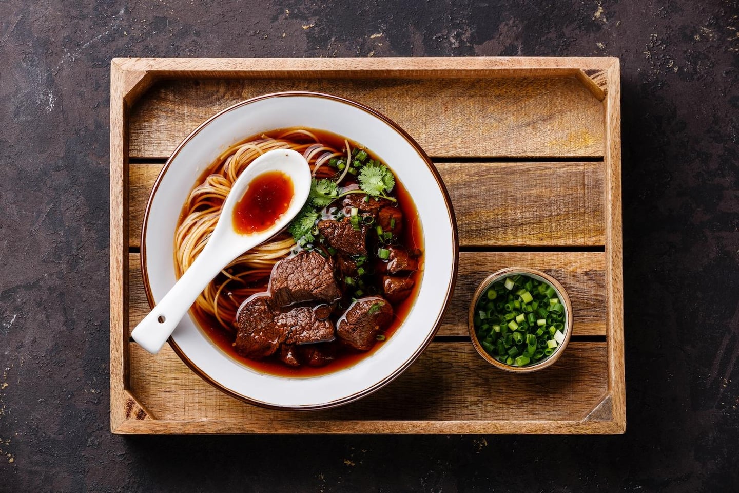 나무 트레이 위 흰 그릇에 담긴 먹음직스러운 소고기 우육면과 잘게 썬 파가 담긴 작은 종지 그릇의 모습입니다.
