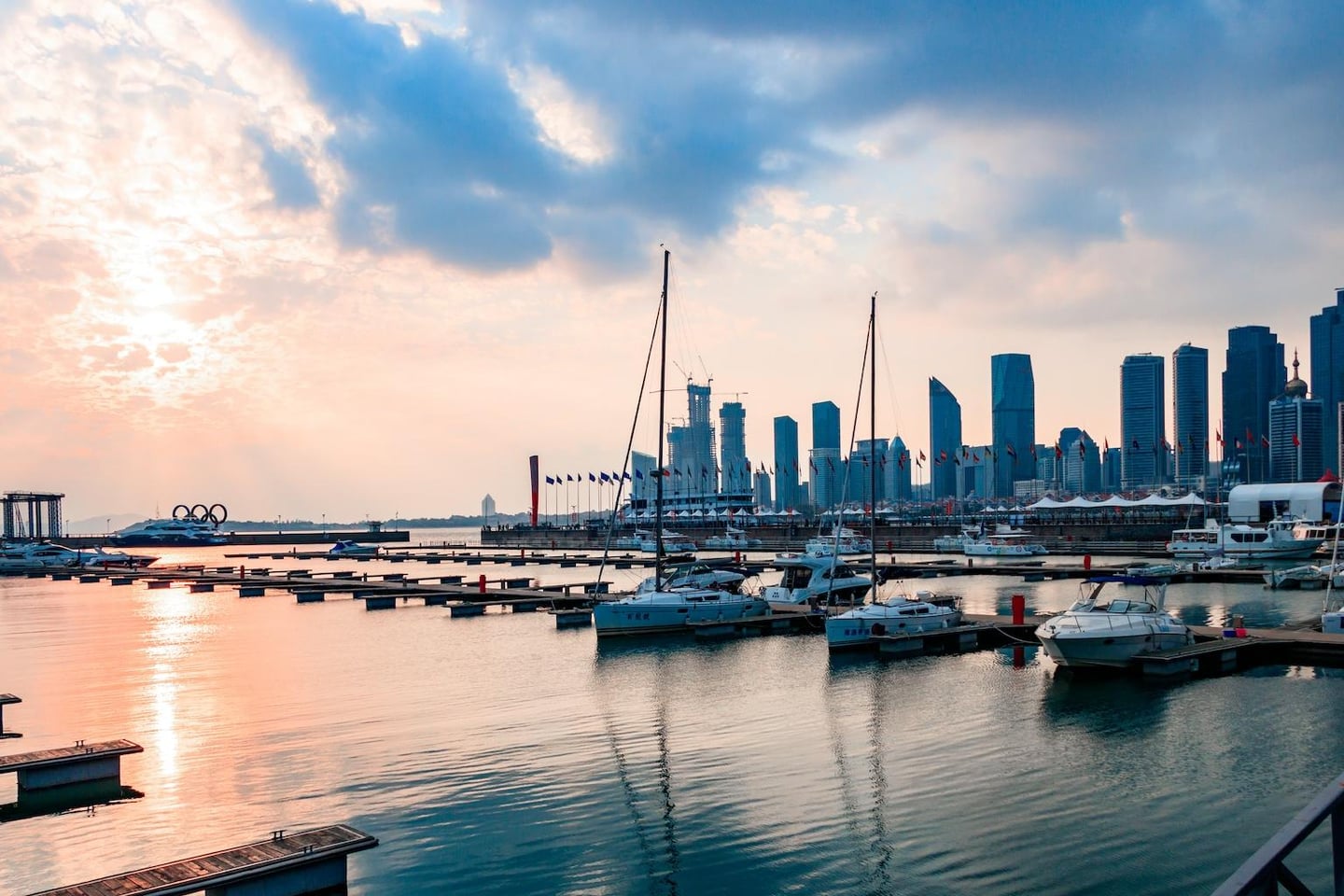 붉게 물든 노을 아래 정박한 요트들과 현대적인 고층 빌딩들이 어우러진 칭다오 올림픽 요트 경기장의 평화로운 저녁 풍경입니다.