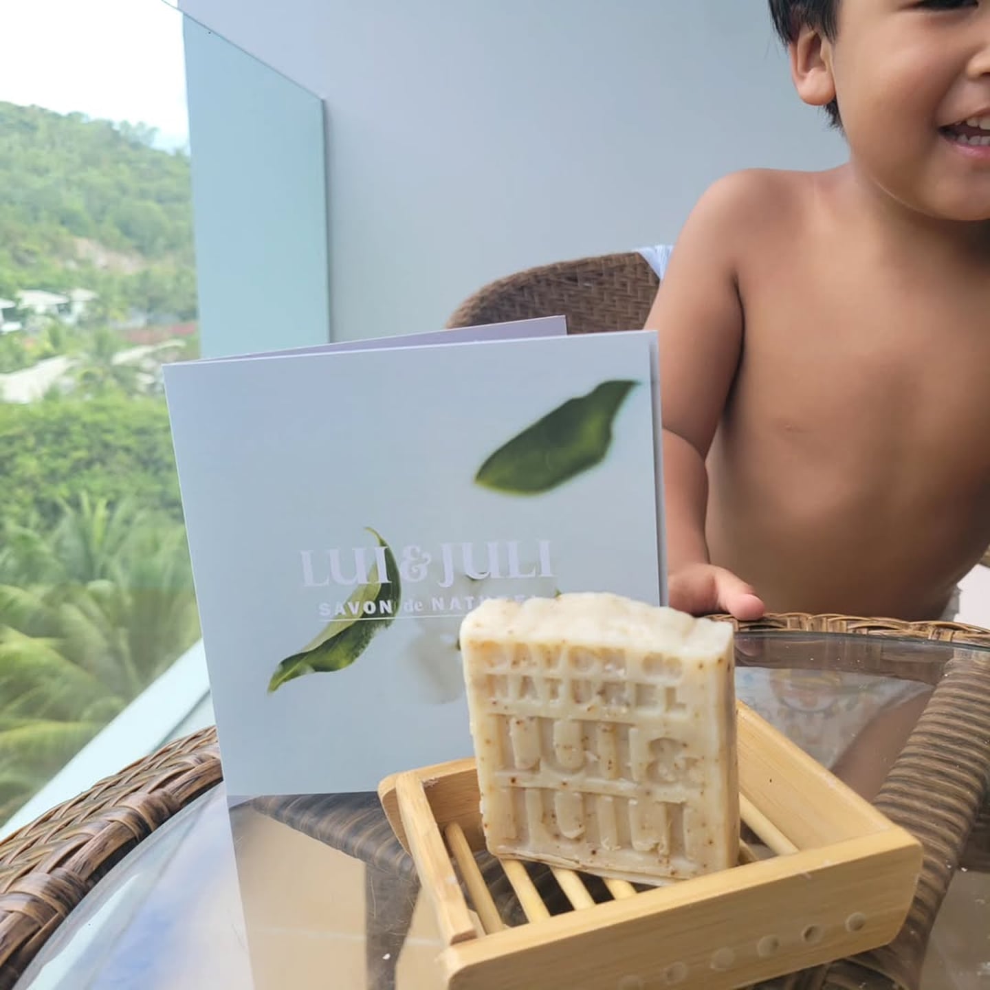 푸른 숲이 내려다보이는 야외 테라스 테이블 위에 놓인 나무 받침대 위의 천연 비누와 브랜드 안내 카드