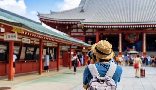밀짚모자와 배낭 차림의 여행자가 카메라를 들고 일본 도쿄 아사쿠사 센소지의 웅장한 붉은 사찰 건물과 활기찬 광장 풍경을 담고 있는 뒷모습입니다.