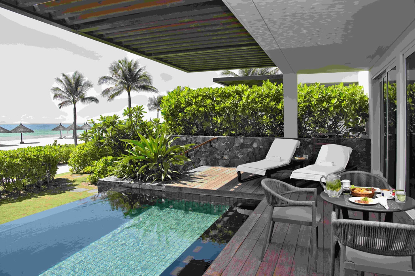 에메랄드빛 바다와 야자수가 보이는 프라이빗 풀 빌라 테라스에 화이트 선베드와 정갈하게 차려진 야외 테이블이 놓여 있는 평화로운 휴양지 전경입니다.