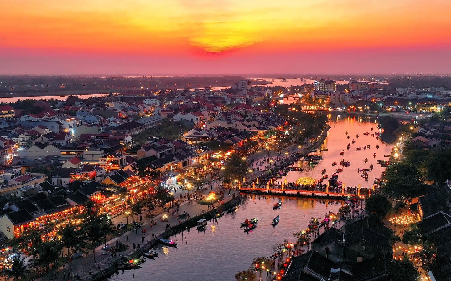 해 질 녘 붉은 노을이 내려앉은 호이안 올드타운의 투본강 위로 나룻배들이 떠다니고 화려한 등불이 켜진 강변 마을의 아름다운 야경입니다.