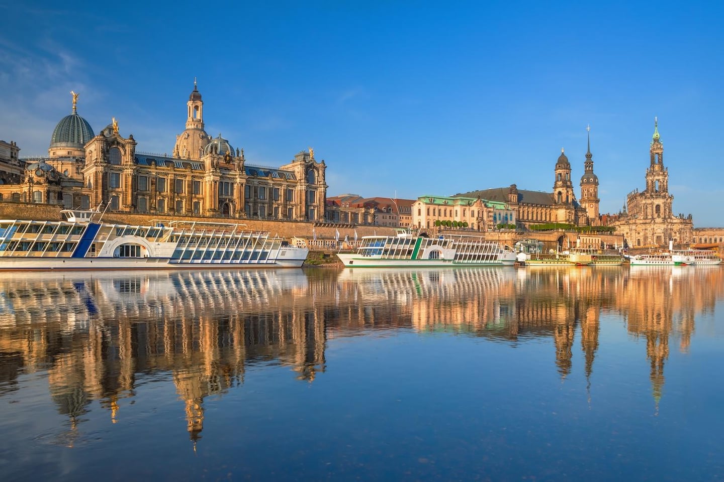 맑은 하늘 아래 드레스덴의 역사적인 건물들과 유람선이 엘베 강물 위로 선명하게 투영된 평화로운 유럽 도시의 전경입니다.