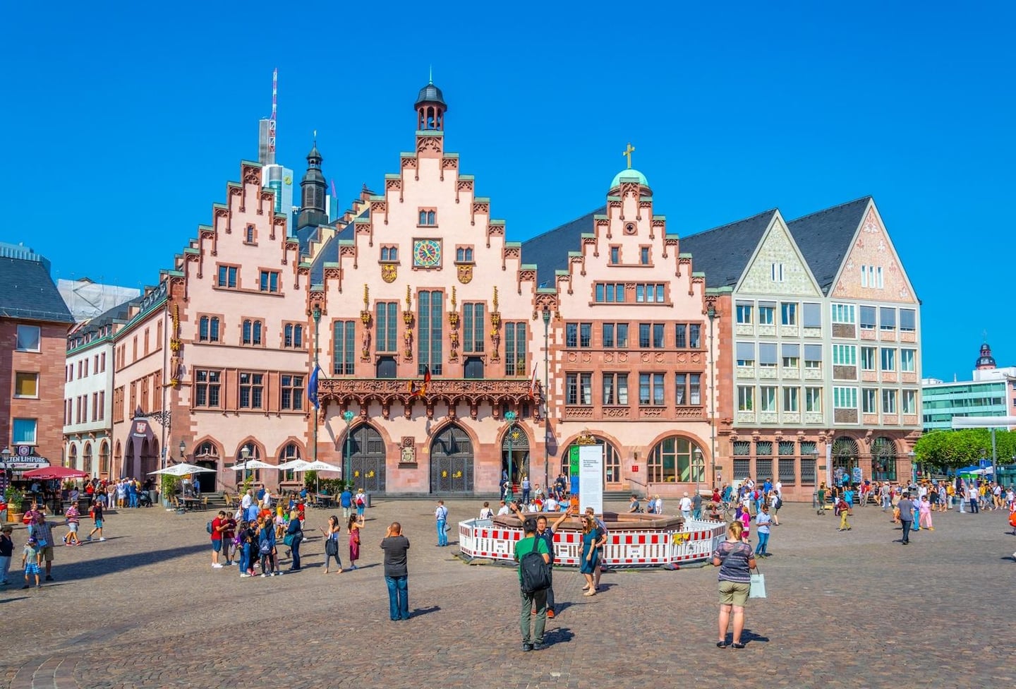 푸른 하늘 아래 독일 프랑크푸르트 뢰머광장의 상징적인 계단식 지붕 건물들과 광장을 거니는 수많은 관광객들의 활기찬 모습입니다.