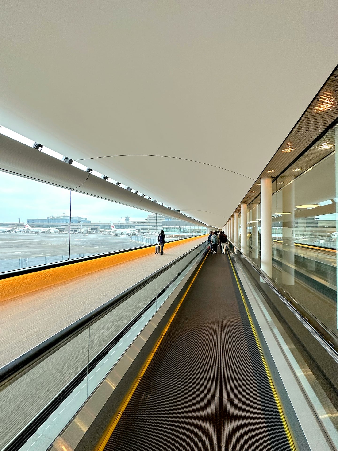 현대적인 공항 터미널 내부에서 무빙워크를 타고 이동하는 여행객들과 창밖으로 보이는 활주로 및 비행기들의 전경입니다.