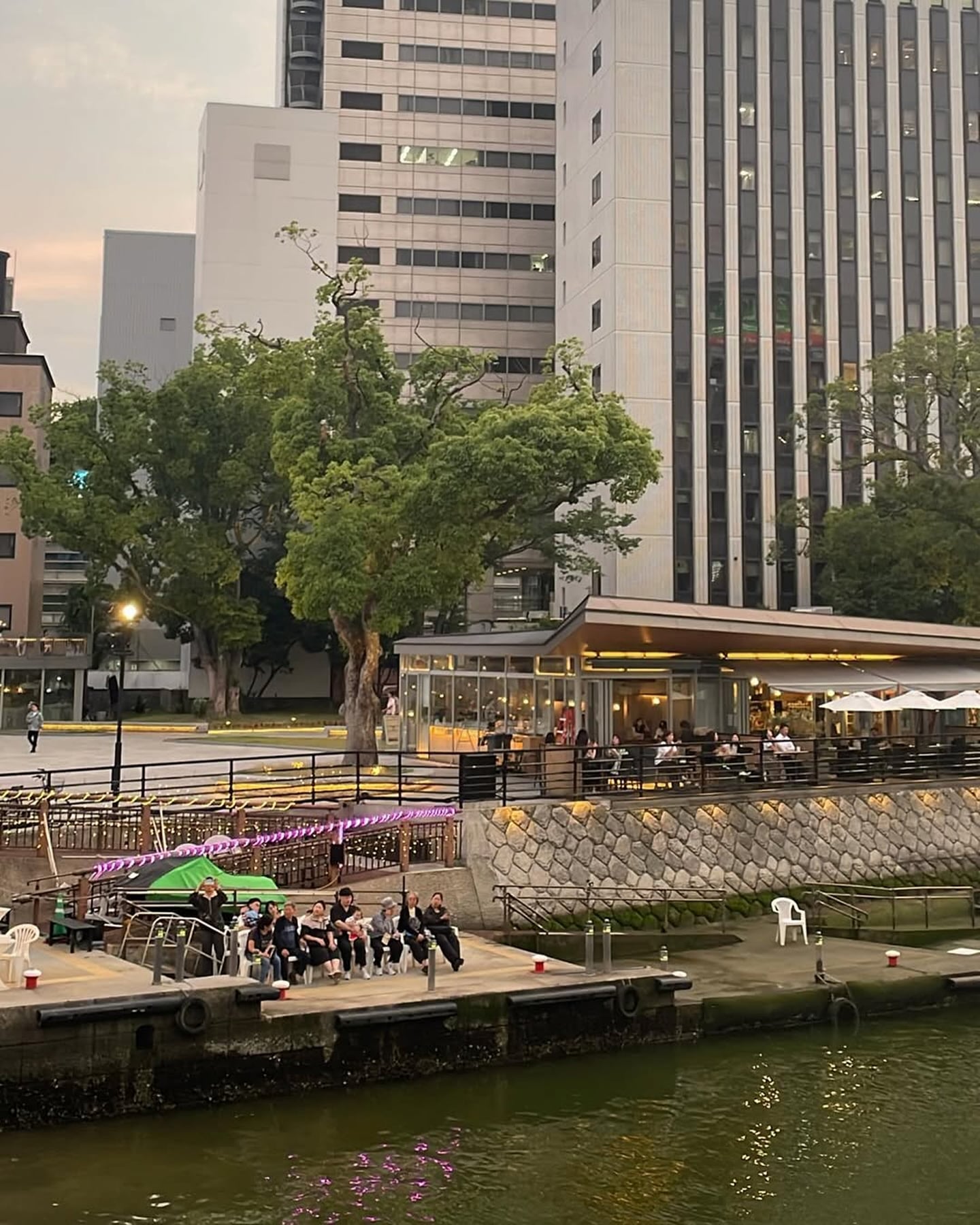 강변의 현대적인 빌딩과 나무들이 어우러진 풍경 속에서 야외 테라스 카페를 이용하거나 선착장에 앉아 여유로운 저녁 시간을 보내는 사람들의 모습입니다.
