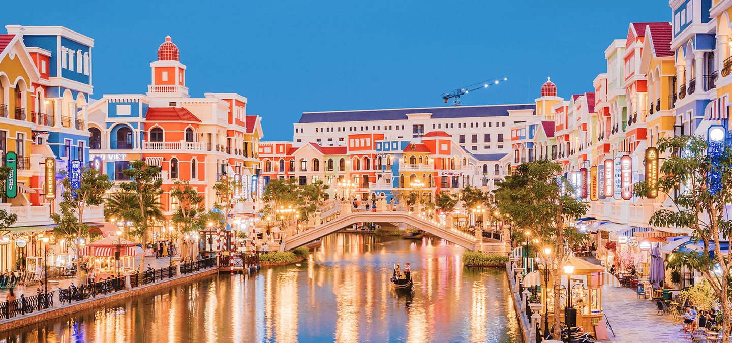 푸른 저녁 하늘 아래 화려한 색채의 건물들과 운하 위를 지나는 곤돌라가 어우러진 베트남 푸꾸옥 그랜드월드의 야경입니다.