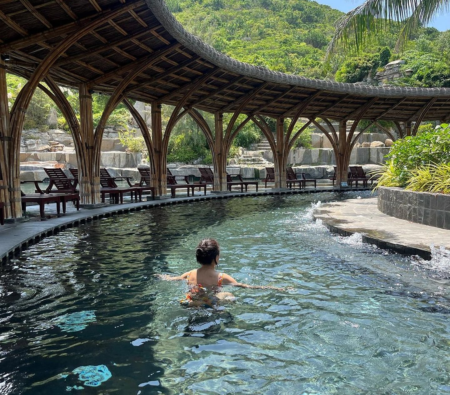 푸른 산이 보이는 야외 온천에서 대나무 아치 지붕과 선베드가 늘어선 수영장을 바라보며 수영을 즐기는 여성의 뒷모습입니다.