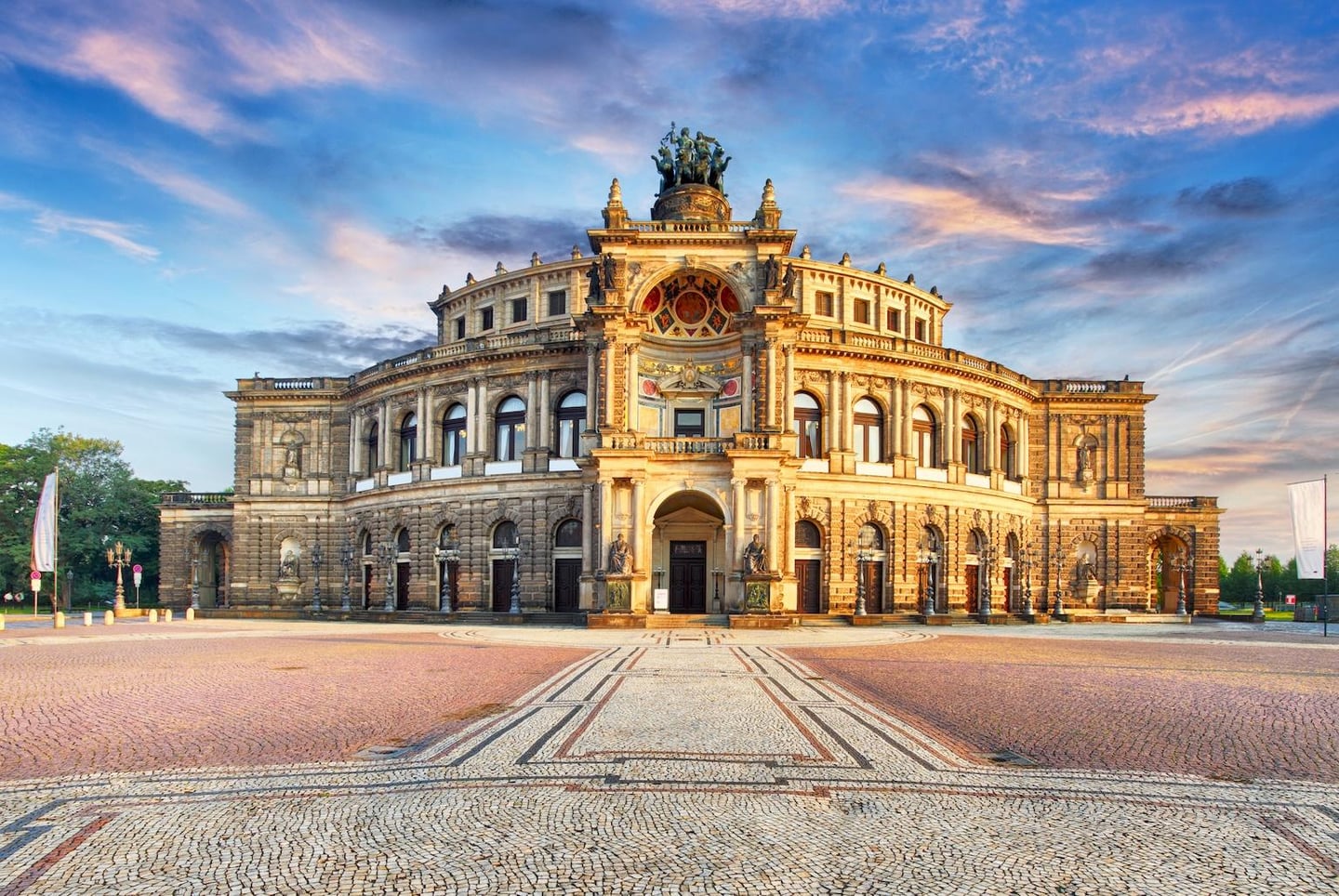 노을빛 하늘 아래 웅장하게 서 있는 드레스덴 젬퍼 오페라 하우스의 화려한 외관과 넓은 돌바닥 광장이 조화를 이루는 풍경입니다.