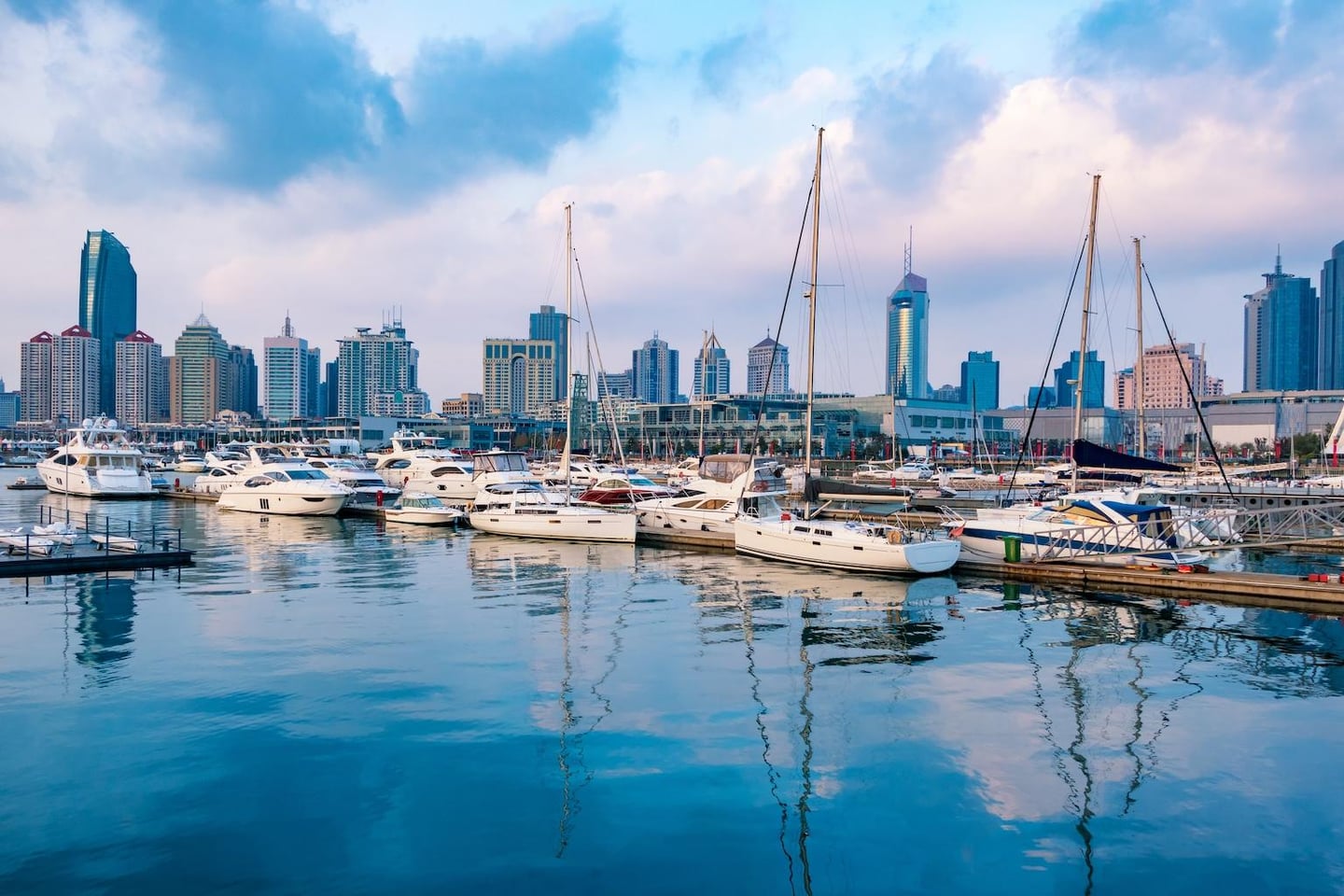 잔잔한 바다 위로 하얀 요트들이 줄지어 정박해 있고 그 뒤로 화려한 도시의 스카이라인이 펼쳐진 항구의 전경입니다.
