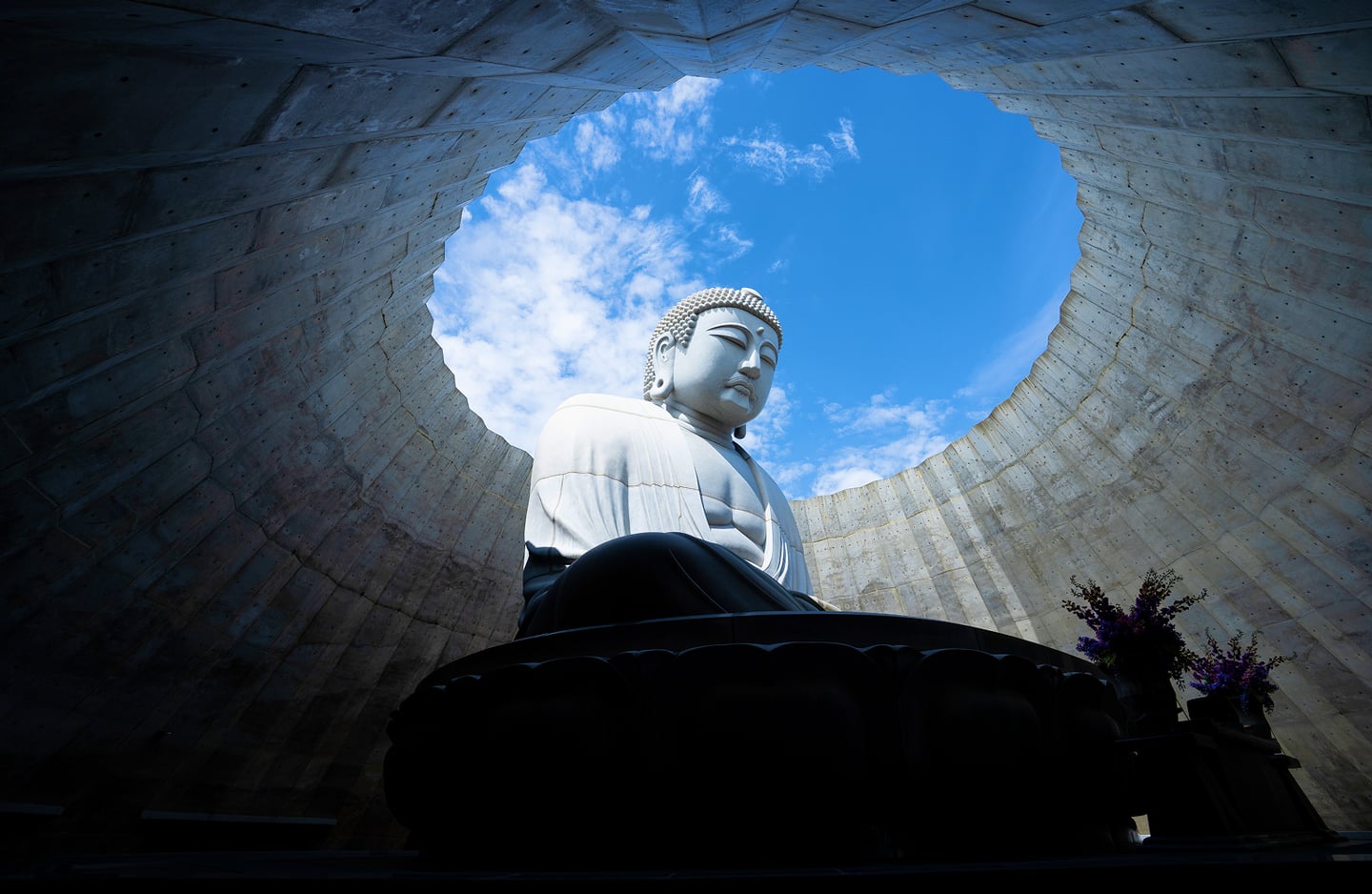 푸른 하늘이 보이는 원형 개구부 아래에 안치된 거대한 석조 불상의 모습으로, 현대적인 건축미와 종교적인 경건함이 조화를 이루는 풍경입니다.