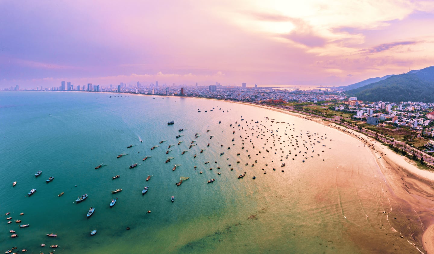 노을 지는 보랏빛 하늘 아래 수많은 어선들이 점점이 떠 있는 다낭 해변과 해안선을 따라 늘어선 도시 건물의 풍경입니다.