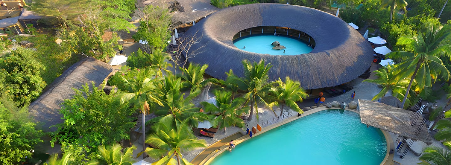 열대 정원 속에 자리 잡은 독특한 원형 건축물과 에메랄드빛 야외 수영장이 어우러진 이국적인 휴양지 리조트의 항공 뷰입니다.