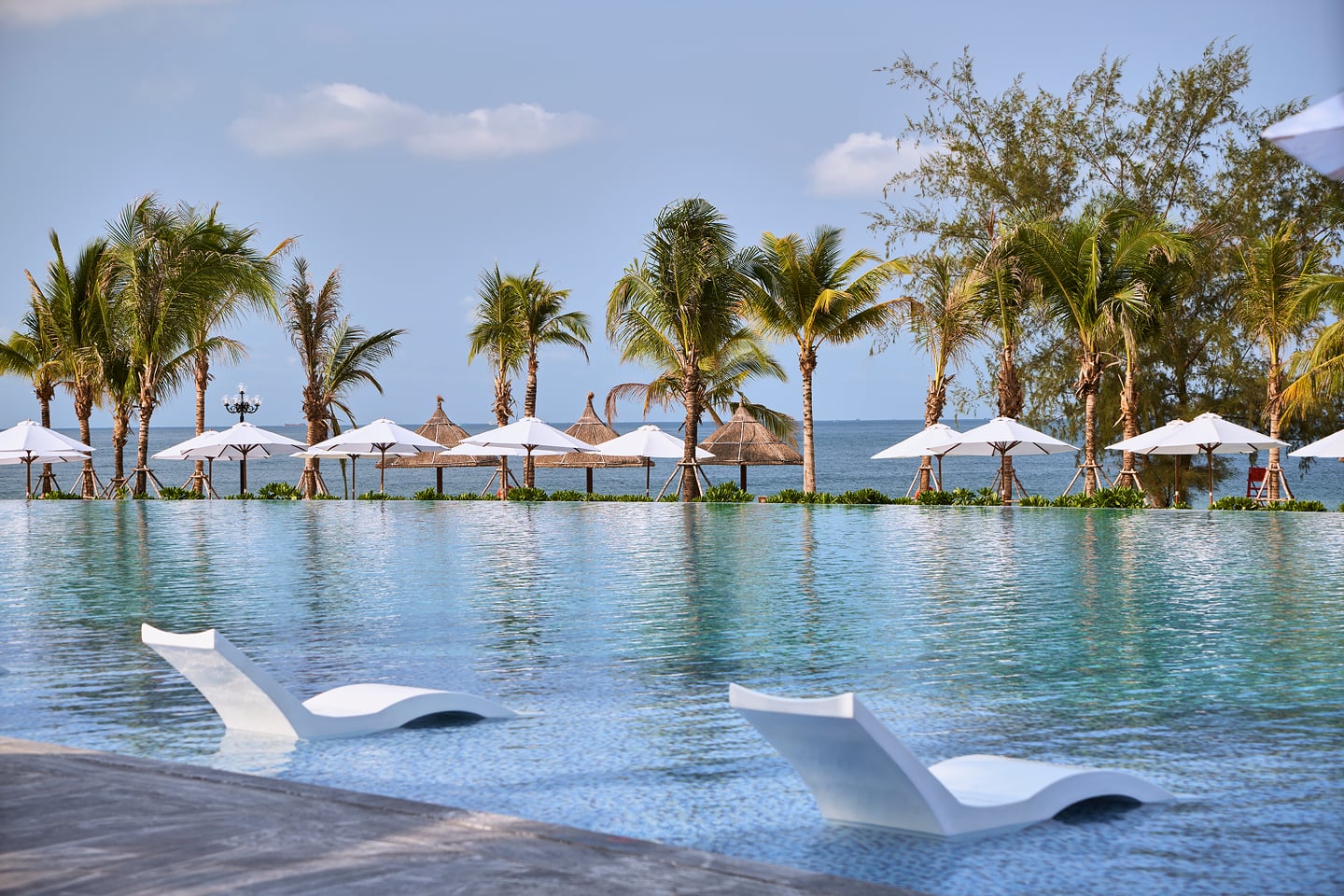 에메랄드빛 인피니티 풀 위에 떠 있는 하얀 선베드와 해변을 따라 늘어선 야자수 및 파라솔이 어우러진 이국적인 리조트 풍경입니다.