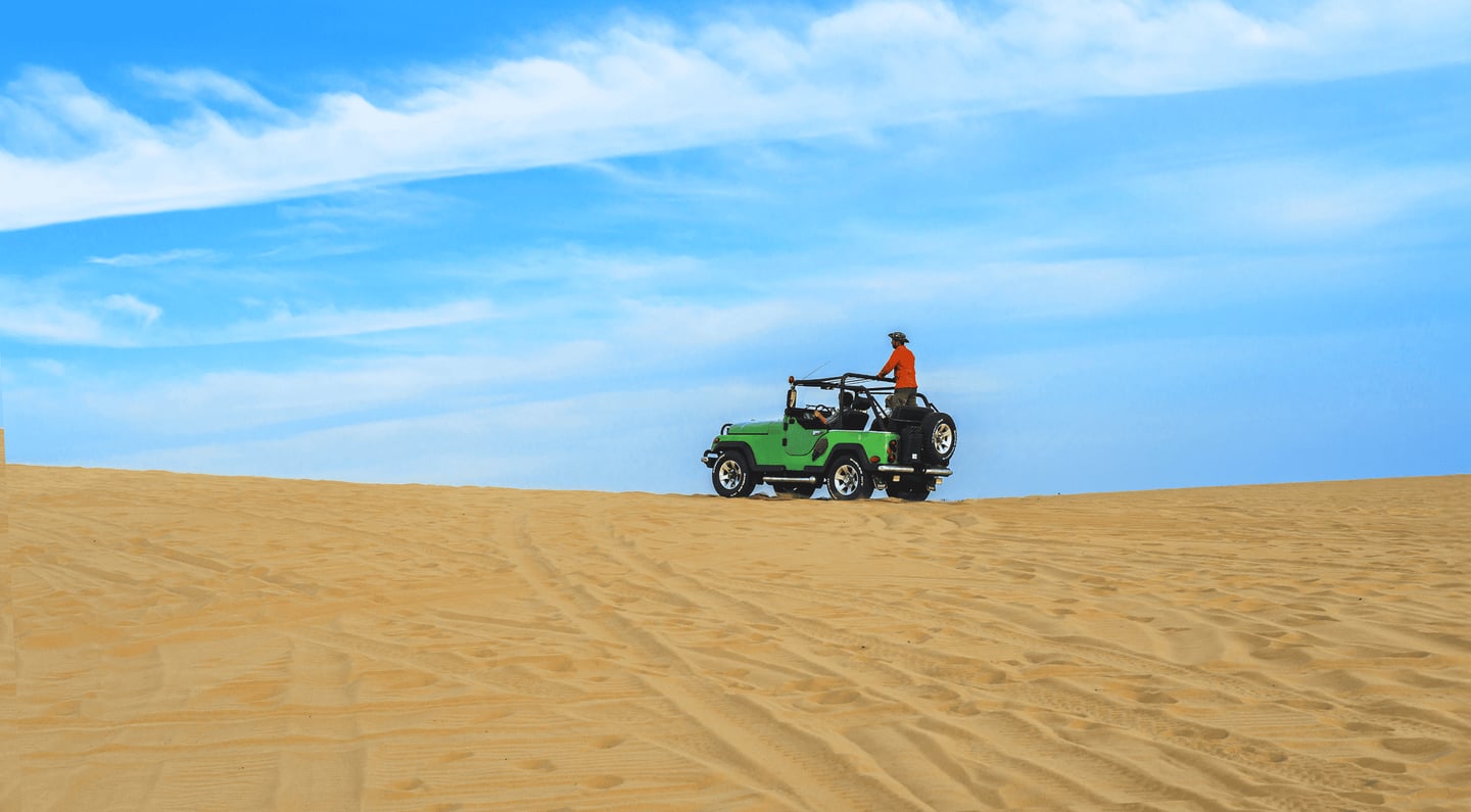 맑고 푸른 하늘 아래 끝없이 펼쳐진 사막의 모래 언덕 위를 주황색 옷을 입은 여행객이 초록색 지프차를 타고 가로지르는 풍경입니다.
