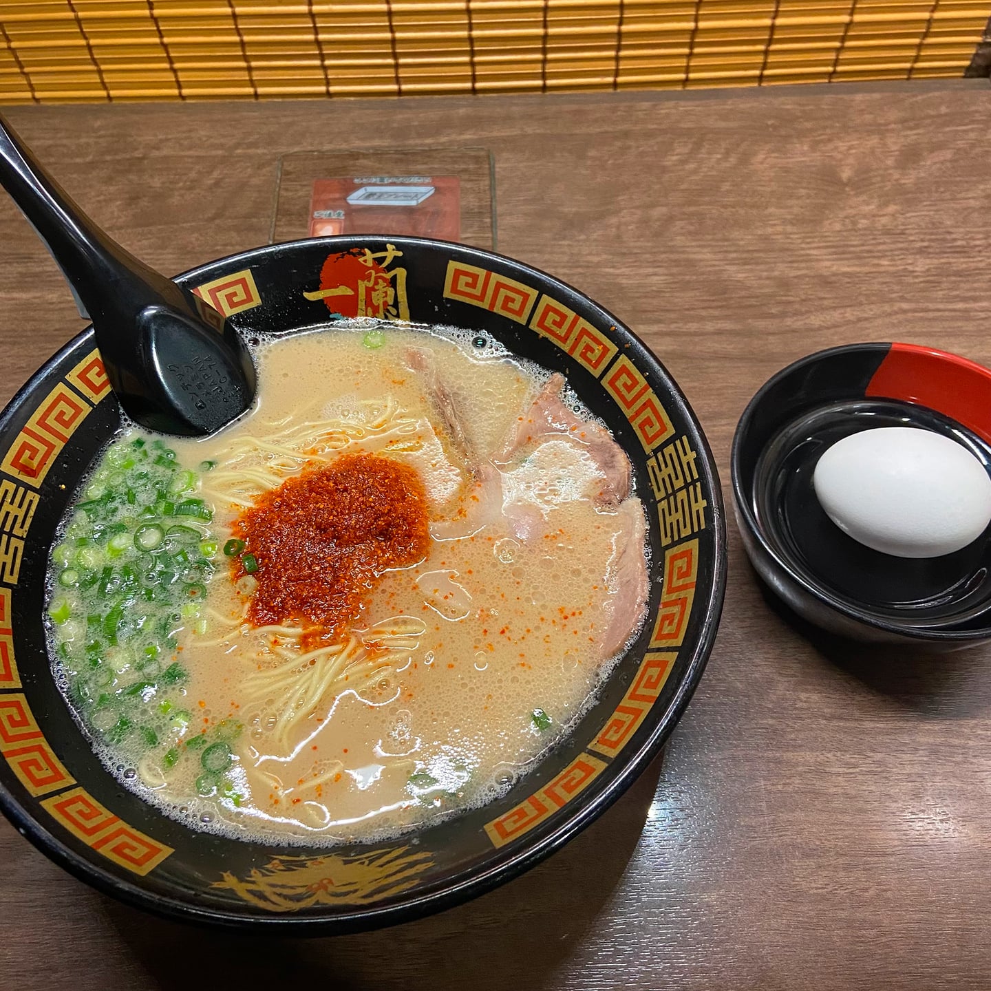 진한 육수와 붉은 양념이 돋보이는 일본 이치란 라멘 한 그릇과 껍질을 까지 않은 삶은 달걀이 나무 테이블 위에 놓인 모습입니다.